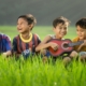 Four boys sitting in tall grass. One with a ukulele and the others singing