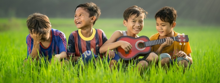 Four boys sitting in tall grass. One with a ukulele and the others singing